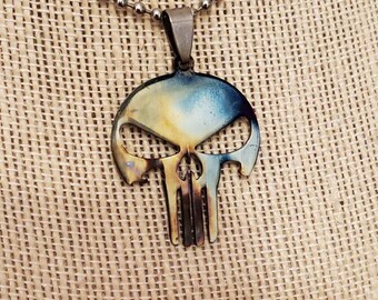 Pode incluir: Um colar de corrente prateada com um pingente de caveira de metal. A caveira é de cor azul escura e dourada com um acabamento texturizado.