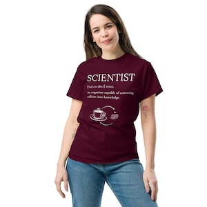 May include: A maroon t-shirt with the word "SCIENTIST" in white, along with a definition and a graphic of a coffee cup and books. The shirt is a crew neck style and is being worn.