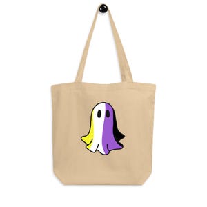 May include: Beige tote bag featuring a cartoon ghost with the non-binary pride flag colors: yellow, white, purple, and black. The bag has a simple design with a long handle for carrying over the shoulder. The ghost is centered on the bag.