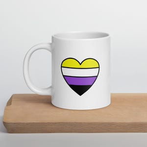 Puede incluir: Taza de cerámica blanca con un diseño en forma de corazón con los colores de la bandera del orgullo no binario: amarillo, morado y negro. La taza tiene un asa curvada y está colocada sobre una superficie de madera. Tamaño estándar.