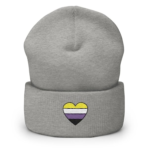 May include: A gray beanie with a heart-shaped embroidered design. The heart features a yellow, white, purple, and black color scheme, representing the non-binary pride flag. The beanie is made of a soft, knitted material.