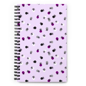 Puede incluir: Un cuaderno con espiral y cubierta lila claro. La cubierta tiene un patrón de pequeñas formas estilizadas negras, blancas, grises y moradas. La encuadernación en espiral del cuaderno es negra.