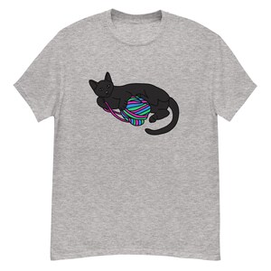 Peut inclure: T-shirt gris clair avec une illustration de chat noir jouant avec une pelote de laine multicolore. La laine est rayée de bleu, vert, rose et violet. Le chat est allongé, les pattes sur la pelote.