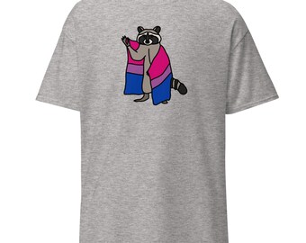 May include: A light gray t-shirt featuring a cartoon raccoon wearing a cape in the colors of the bisexual pride flag: pink, purple, and blue. The raccoon is standing with one paw raised.