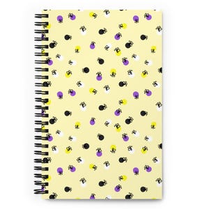 May include: A spiral-bound notebook with a pale yellow cover. The cover is patterned with small, stylized black, white, yellow, and purple shapes. The spiral binding is black.