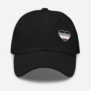 May include: Black baseball cap with a heart-shaped embroidered design. The heart features a striped pattern in white, gray, and purple, with a black outline. The cap has a curved brim and a button on top.