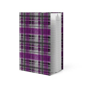 Puede incluir: Un cuaderno de tapa dura con un patrón de cuadros morado, gris y negro. El cuaderno está abierto, revelando páginas blancas rayadas. El diseño de la cubierta presenta un efecto texturizado, similar a una pincelada.