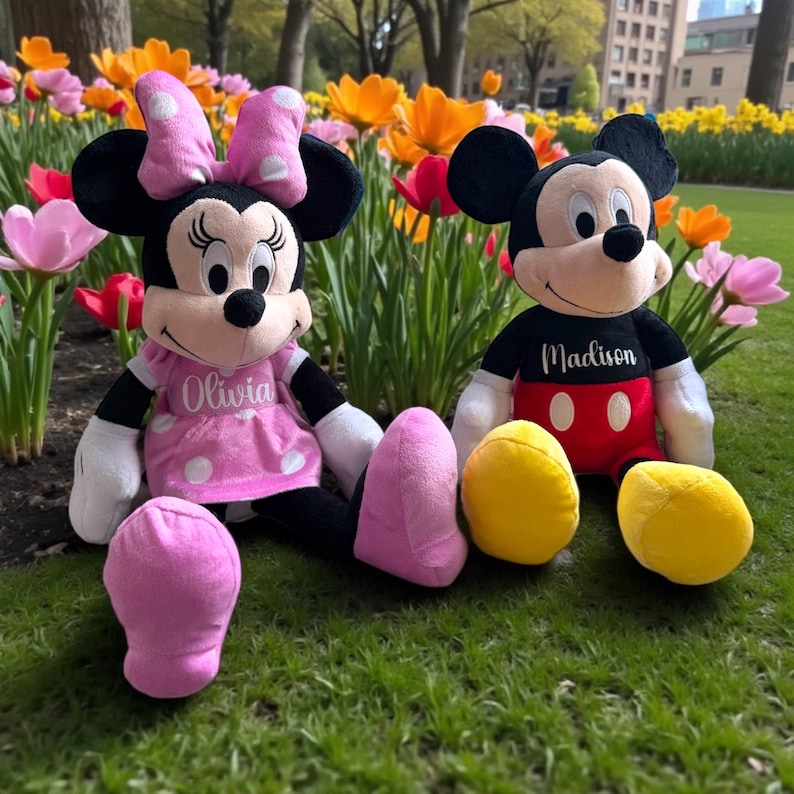 Puede incluir: Dos mu&ntilde;ecos de peluche de los personajes de Disney, Minnie Mouse y Mickey Mouse, sentados sobre hierba frente a flores coloridas. Minnie lleva un vestido rosa con lunares blancos y el nombre "Olivia". Mickey lleva una camisa negra con el nombre "Madison".