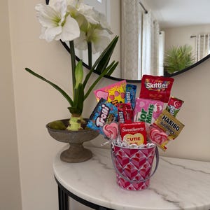 Puede incluir: Un arreglo decorativo con un cubo rosa lleno de caramelos y piruletas. El cubo tiene un patr&oacute;n de corazones. Tambi&eacute;n hay una planta en maceta con flores blancas y hojas verdes.