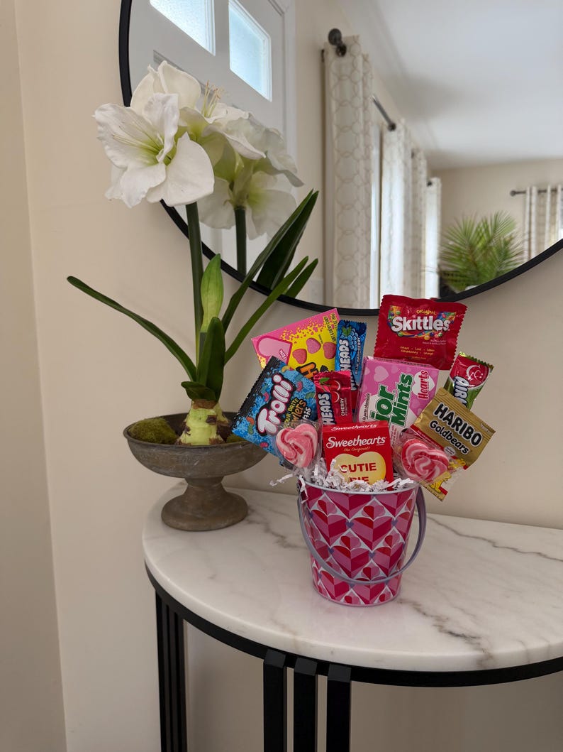 Puede incluir: Un arreglo decorativo con un cubo rosa lleno de caramelos y piruletas. El cubo tiene un patr&oacute;n de corazones. Tambi&eacute;n hay una planta en maceta con flores blancas y hojas verdes.