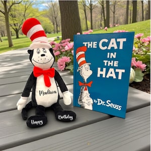 May include: A plush Cat in the Hat doll with a red and white striped hat and bow tie, next to a copy of "The Cat in the Hat" book by Dr. Seuss. The doll has the name "Madison" on its chest and "Happy Birthday" on its feet.