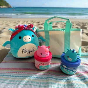 May include: A teal plush dragon toy with a red patterned headband and the name Abigail printed in black, a cream canvas tote bag with mint green handles, and two plastic jars topped with small pink and teal dragon figurines.