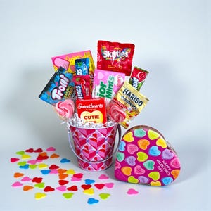 Puede incluir: Un ramo de caramelos de San Valent&iacute;n en un cubo rosa con un patr&oacute;n de corazones, lleno de varios caramelos como Skittles, Haribo Goldbears y Sweethearts. Una caja en forma de coraz&oacute;n con corazones de caramelo est&aacute; al lado del cubo.