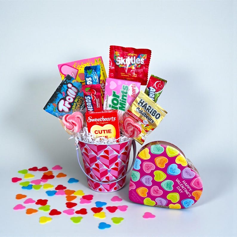 Puede incluir: Un ramo de caramelos de San Valent&iacute;n en un cubo rosa con un patr&oacute;n de corazones, lleno de varios caramelos como Skittles, Haribo Goldbears y Sweethearts. Una caja en forma de coraz&oacute;n con corazones de caramelo est&aacute; al lado del cubo.
