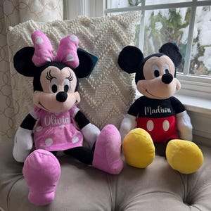 Puede incluir: Dos mu&ntilde;ecos de peluche de los personajes de Disney, Minnie Mouse y Mickey Mouse, est&aacute;n uno al lado del otro. Minnie lleva un vestido y un lazo rosas con lunares blancos. Mickey tiene una camisa negra y pantalones cortos rojos. C&iacute;rculos de peluche amarillos est&aacute;n delante.
