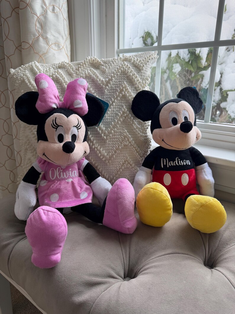 Puede incluir: Dos mu&ntilde;ecos de peluche de los personajes de Disney, Minnie Mouse y Mickey Mouse, est&aacute;n uno al lado del otro. Minnie lleva un vestido y un lazo rosas con lunares blancos. Mickey tiene una camisa negra y pantalones cortos rojos. C&iacute;rculos de peluche amarillos est&aacute;n delante.
