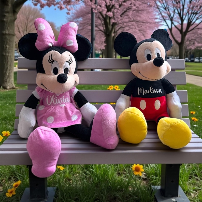 Puede incluir: Dos mu&ntilde;ecos de peluche de los personajes de Disney, Minnie Mouse y Mickey Mouse, sentados en un banco del parque. Minnie lleva un vestido rosa con lunares blancos y un lazo. Mickey lleva una camisa negra con un nombre y pantalones cortos rojos. Hay cojines amarillos cerca.