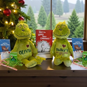 May include: Two green plush Grinch dolls with embroidered names Olivia and Hailey's 1st Christmas 2025, positioned beside a How the Grinch Stole Christmas book, two Swiss Miss milk chocolate cards, two small boxes of Hershey's Kisses, and candy canes.