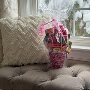 Puede incluir: Un cubo de caramelos rosa y blanco con estampado de corazones, lleno de varios caramelos, envuelto en celof&aacute;n y atado con una cinta rosa. El cubo est&aacute; sobre un coj&iacute;n acolchado junto a una almohada blanca estampada, con una ventana nevada al fondo.