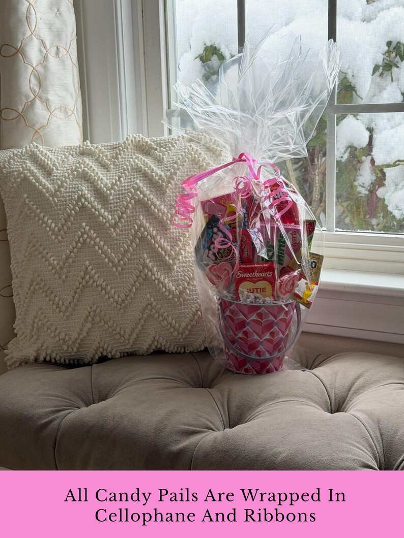 Puede incluir: Un cubo de caramelos rosa y blanco con estampado de corazones, lleno de varios caramelos, envuelto en celof&aacute;n y atado con una cinta rosa. El cubo est&aacute; sobre un coj&iacute;n acolchado junto a una almohada blanca estampada, con una ventana nevada al fondo.