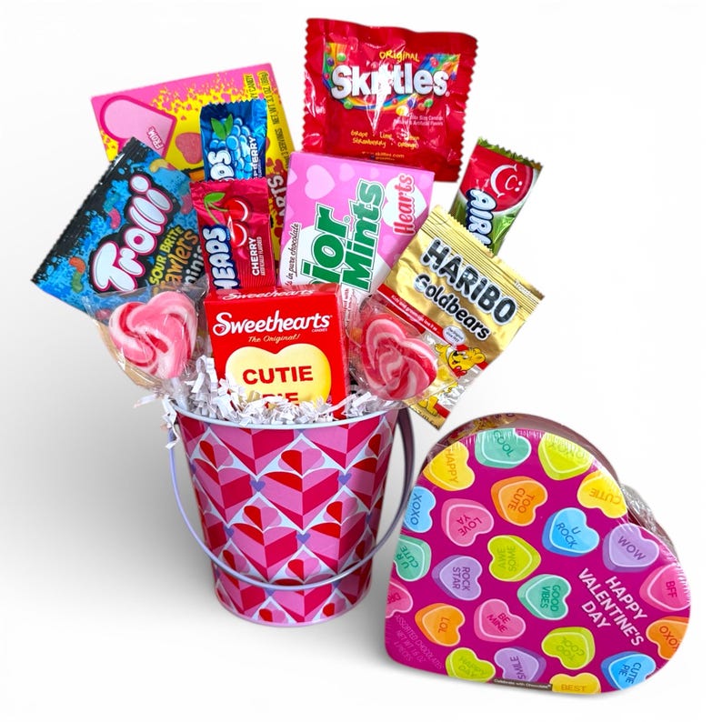 Puede incluir: Un arreglo de caramelos de San Valent&iacute;n en un cubo rosa con un patr&oacute;n de corazones, lleno de varios caramelos y piruletas. Una caja en forma de coraz&oacute;n con corazones de caramelo coloridos y el texto "Happy Valentine's Day" est&aacute; al lado del cubo.