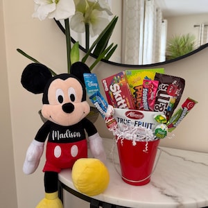 Puede incluir: Un peluche de Mickey Mouse con el nombre "Madison" en su camisa est&aacute; junto a un cubo rojo lleno de caramelos. El cubo est&aacute; sobre una mesa con tapa de m&aacute;rmol, con una pelota amarilla y flores blancas en el fondo.