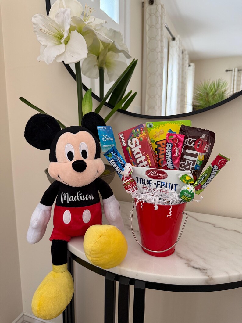 Puede incluir: Un peluche de Mickey Mouse con el nombre "Madison" en su camisa est&aacute; junto a un cubo rojo lleno de caramelos. El cubo est&aacute; sobre una mesa con tapa de m&aacute;rmol, con una pelota amarilla y flores blancas en el fondo.