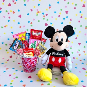 Puede incluir: Un peluche de Mickey Mouse con el nombre "Madison" en la camisa est&aacute; junto a un peque&ntilde;o cubo lleno de caramelos. El cubo tiene un patr&oacute;n de corazones rosas y rojos. Las marcas de caramelos incluyen Skittles, Trolli y Haribo. El fondo tiene un patr&oacute;n de corazones coloridos.