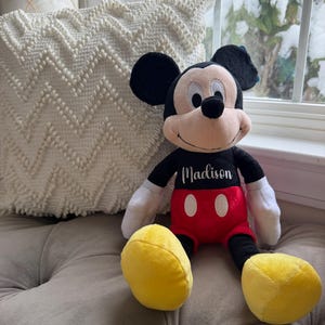 Puede incluir: Mu&ntilde;eco de peluche de Mickey Mouse con orejas negras, cuerpo negro, pantalones cortos rojos y pies amarillos. El mu&ntilde;eco tiene guantes blancos y el nombre "Madison" bordado en el pecho. El mu&ntilde;eco est&aacute; sentado sobre una superficie gris.