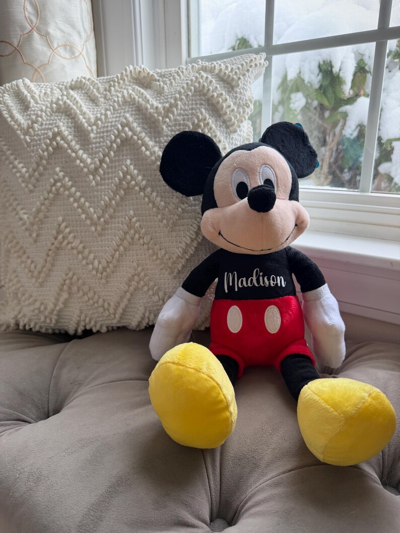 Puede incluir: Mu&ntilde;eco de peluche de Mickey Mouse con orejas negras, cuerpo negro, pantalones cortos rojos y pies amarillos. El mu&ntilde;eco tiene guantes blancos y el nombre "Madison" bordado en el pecho. El mu&ntilde;eco est&aacute; sentado sobre una superficie gris.