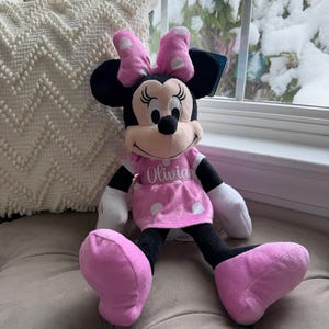 Puede incluir: Una mu&ntilde;eca de peluche de Minnie Mouse con un vestido y lazo rosas, lunares blancos y el nombre "Olivia" bordado en el vestido. La mu&ntilde;eca tiene orejas, brazos y piernas negros y guantes blancos. La mu&ntilde;eca est&aacute; sentada en un coj&iacute;n gris.