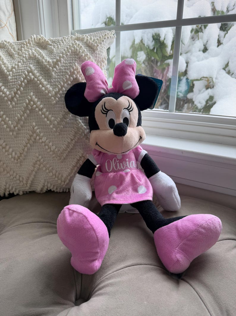 Puede incluir: Una mu&ntilde;eca de peluche de Minnie Mouse con un vestido y lazo rosas, lunares blancos y el nombre "Olivia" bordado en el vestido. La mu&ntilde;eca tiene orejas, brazos y piernas negros y guantes blancos. La mu&ntilde;eca est&aacute; sentada en un coj&iacute;n gris.
