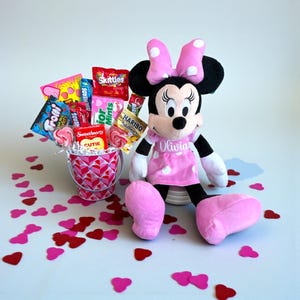Puede incluir: Un peluche de Minnie Mouse con un vestido rosa con lunares, con el nombre "Olivia" bordado. Un cubo lleno de caramelos y confeti en forma de coraz&oacute;n est&aacute; junto al peluche. Los caramelos incluyen piruletas, Skittles y Haribo.