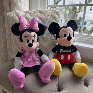 Puede incluir: Dos mu&ntilde;ecos de peluche de los personajes de Disney, Minnie Mouse y Mickey Mouse, sentados uno al lado del otro. Minnie lleva un vestido rosa con lunares blancos y un lazo a juego. Mickey lleva pantalones cortos rojos. Ambos mu&ntilde;ecos tienen nombres personalizados en sus camisas.