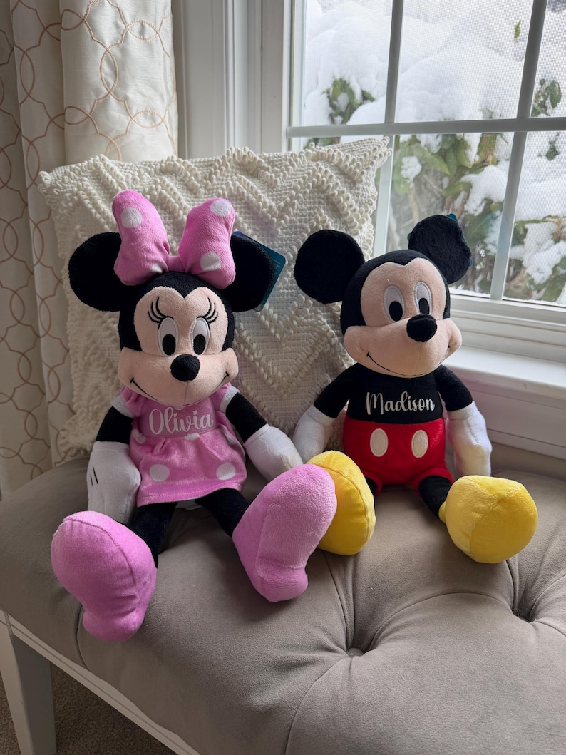 Puede incluir: Dos mu&ntilde;ecos de peluche de los personajes de Disney, Minnie Mouse y Mickey Mouse, sentados uno al lado del otro. Minnie lleva un vestido rosa con lunares blancos y un lazo a juego. Mickey lleva pantalones cortos rojos. Ambos mu&ntilde;ecos tienen nombres personalizados en sus camisas.