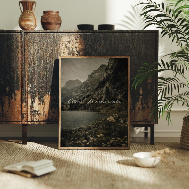 May include: A framed landscape print featuring a mountain range and a lake, with the text "Climb the mountain." The print is in a wooden frame and rests on a woven rug. Decorative vases and bowls are on a dark wooden cabinet.