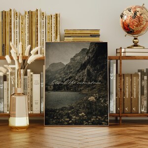 May include: A framed landscape print featuring a mountain range and a lake, with the text "Climb the mountain." The print is displayed on a wooden surface, surrounded by books, a globe, and decorative items. The color palette is muted, with sepia tones.