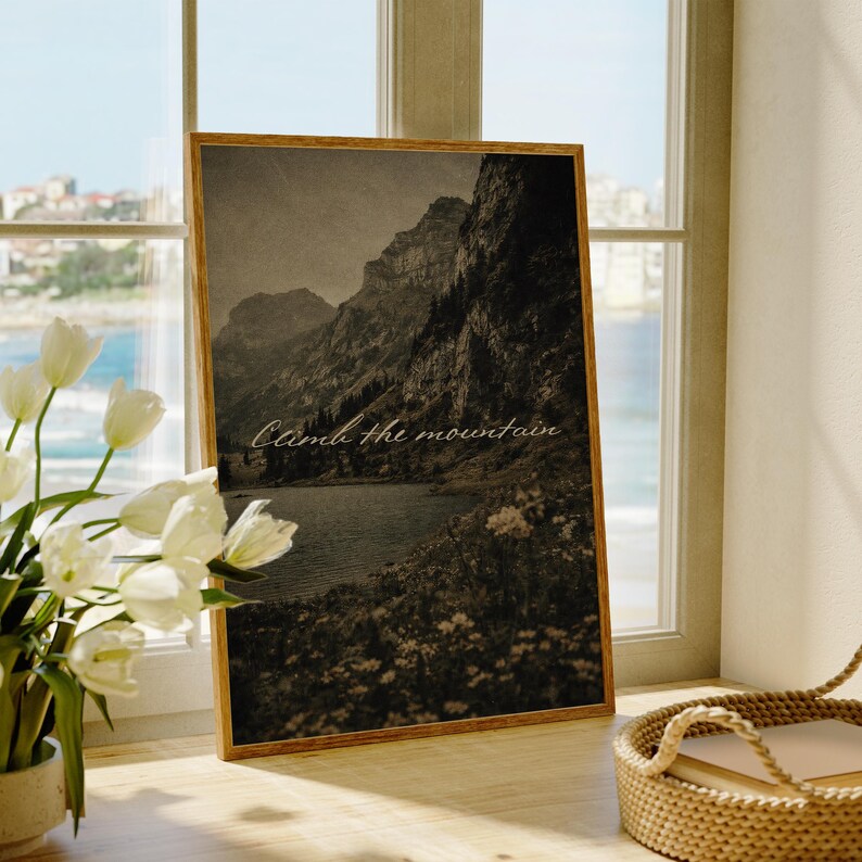May include: A framed sepia-toned print featuring a mountain landscape with a lake and the text "Climb the mountain." The artwork is displayed on a windowsill, with white flowers in a vase to the left and a woven basket to the right.