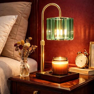 May include: A bedside table lamp with a gold-colored arched frame and a green, faceted glass shade. A lit candle in a glass jar sits on a wooden base, along with a small vase of dried flowers and a vintage alarm clock.