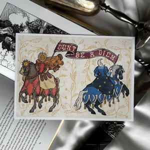 May include: Greeting card with medieval illustrations of knights on horseback. One knight is adorned with a sun, the other with a crescent moon. A banner above reads "DON'T BE A DICK" in burgundy lettering. The background has a floral design.