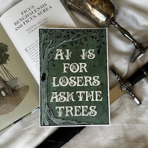 K&ouml;nnte beinhalten: Ein gr&uuml;ner und wei&szlig;er Kunstdruck mit dem Text "AI IS FOR LOSERS ASK THE TREES" wird angezeigt. Der Druck zeigt eine Baumillustration. Ein Buch und ein dekorativer Kelch sind ebenfalls sichtbar.