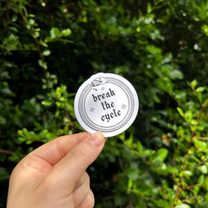 May include: A circular white sticker featuring a black line art illustration of an ouroboros snake eating its own tail. The center contains the black text "break the cycle" in a gothic font, flanked by two small four-pointed star symbols.