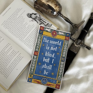 Op de afbeelding: Een open boek, een decoratieve kaart met de tekst "the world is not kind, but I shall be", een zilveren beker en een zwart zwaard met een zilveren handvat liggen op een wit oppervlak.
