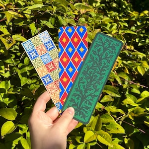 May include: Three rectangular bookmarks with distinct designs. One features a grid of blue, red, and cream floral tiles. Another displays a blue diamond pattern with yellow stars. The third is a solid green with a dark, intricate leaf motif.