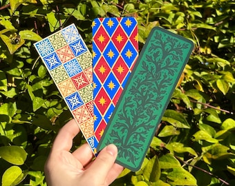May include: Three rectangular bookmarks with distinct designs. One features a grid of blue, red, and cream floral tiles. Another displays a blue diamond pattern with yellow stars. The third is a solid green with a dark, intricate leaf motif.