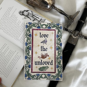 Könnte beinhalten: Dekorative Grußkarte mit dem Text "love the unloved" im mittelalterlichen Stil, mit Illustrationen einer Taube, einer Maus und eines Frosches. Die Karte hat einen floralen Rahmen und Sternakzente.
