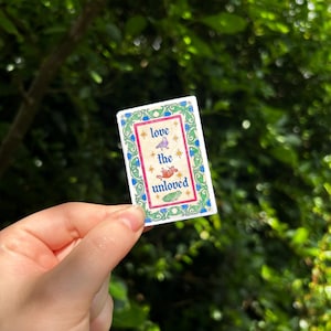 May include: A rectangular sticker with a decorative green and blue floral border. The center features a cream background with the text "love the unloved" in blue, surrounded by small gold stars and illustrations of a bird, a rat, and a frog.
