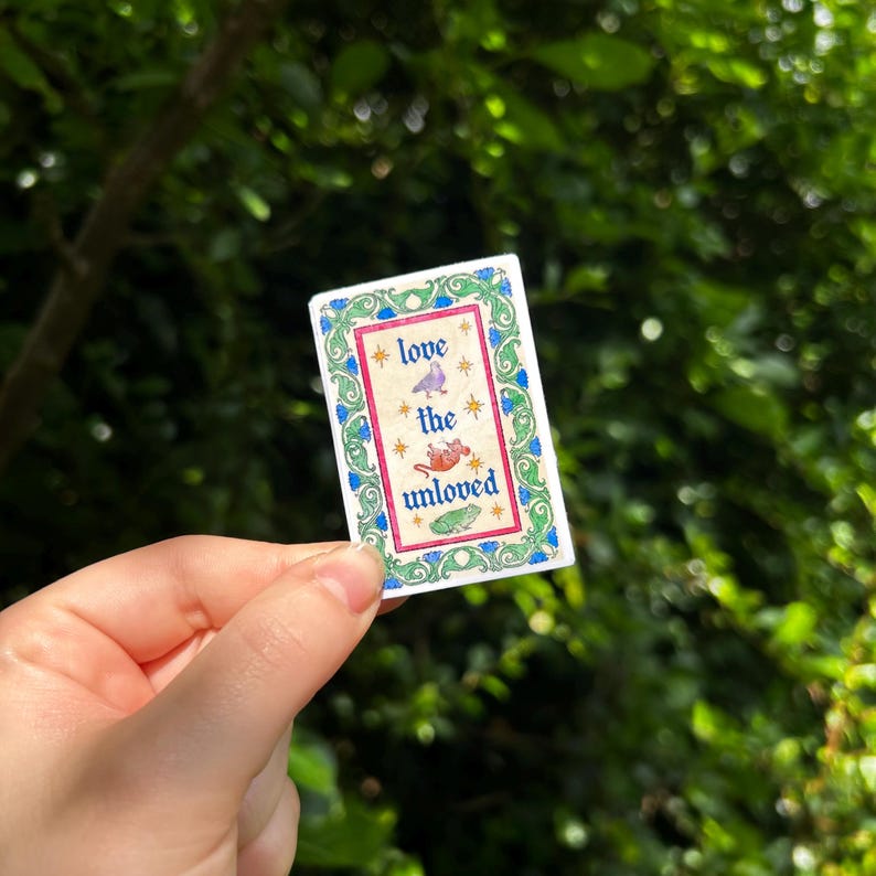May include: A rectangular sticker with a decorative green and blue floral border. The center features a cream background with the text "love the unloved" in blue, surrounded by small gold stars and illustrations of a bird, a rat, and a frog.
