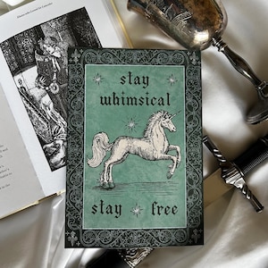 May include: A greeting card featuring a white unicorn illustration on a green background. The card has the words "stay whimsical" and "stay free" in a gothic font. The card is framed by a decorative border.