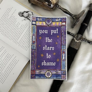 May include: A purple art print with the text "you put the stars to shame" in a decorative frame. The print is surrounded by a book, a sword, and a silver goblet.